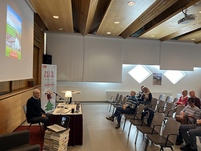 Edwin Klausmann sitzt vor der Leinwand mit eingeblendetem Buchcover, die etwa 10 Zuschauer sitzen auf mehrere Reihen verteilt